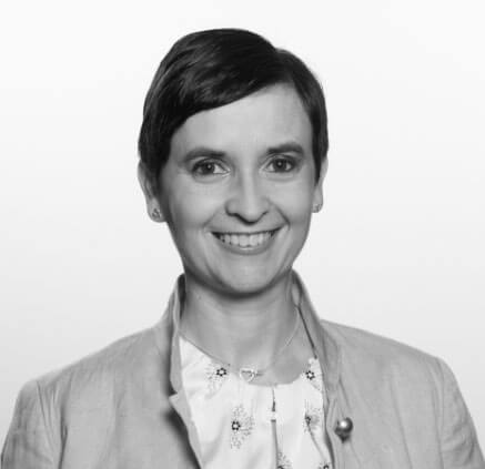 Eine Frau mit kurzen dunklen Haaren lächelt in die Kamera. Sie trägt eine helle Jacke über einer gemusterten Bluse und eine herzförmige Halskette. Der Hintergrund ist schlicht und hell.